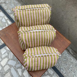 May include: A set of three rectangular fabric pouches in varying sizes, each featuring a vertical striped pattern in olive green, white, and thin pink lines. Each pouch has a white zipper closure with a silver-toned circular metal pull tab.