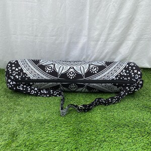 May include: Black and white yoga mat bag with a mandala design. The bag has a zipper closure and an adjustable shoulder strap. The bag is cylindrical and made of a patterned fabric. The bag is designed to carry a yoga mat.