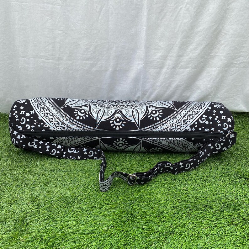 May include: Black and white yoga mat bag with a mandala design. The bag has a zipper closure and an adjustable shoulder strap. The bag is cylindrical and made of a patterned fabric. The bag is designed to carry a yoga mat.