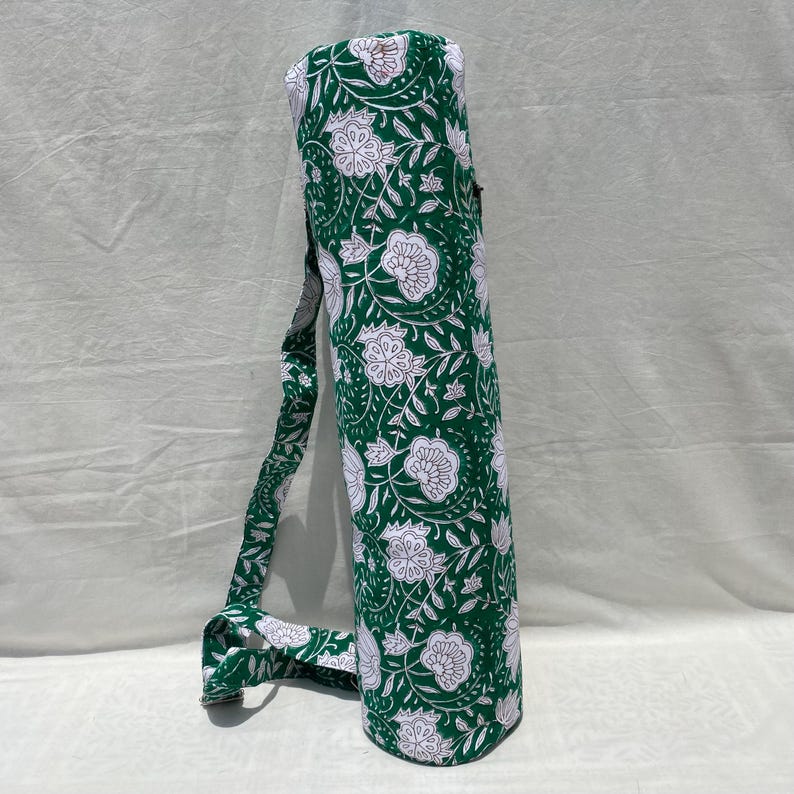 K&ouml;nnte beinhalten: Gr&uuml;ne Yogamatten-Tasche mit wei&szlig;em Blumenmuster und passendem Schultergurt. Die zylindrische Tasche ist f&uuml;r den Transport einer zusammengerollten Yogamatte konzipiert. Der Stoff hat ein sich wiederholendes Muster aus stilisierten Blumen und Bl&auml;ttern.