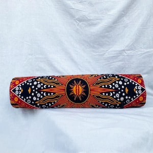 Puede incluir: Un cojín cilíndrico con un vibrante diseño de sol y celestial en rojo, naranja, negro y blanco. El diseño presenta un motivo central de sol, rodeado de lunas crecientes y estrellas. El cojín se utiliza probablemente para yoga o meditación.