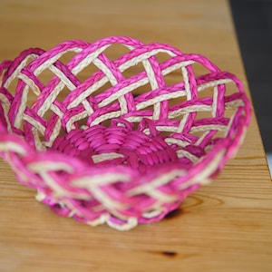 Puede incluir: Una cesta redonda tejida de paja en color rosa y blanco con un patrón decorativo.