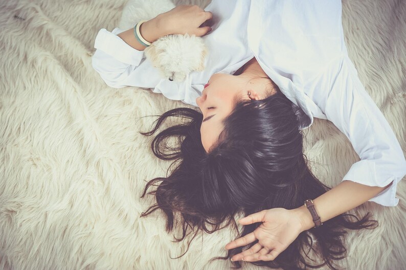 May include: A woman with long dark hair is sleeping on a white fluffy rug. She is wearing a white button-down shirt and has a small white dog in her arms.