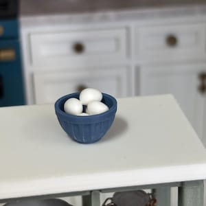May include: A miniature blue bowl filled with white eggs sits on a white countertop. The bowl has a ribbed design. The background shows a miniature kitchen with white cabinets and a blue stove.