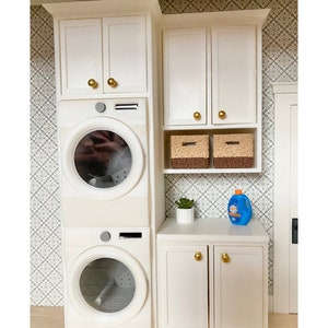 May include: White laundry room cabinet kit with gold knobs. The kit includes upper cabinets, lower cabinets, and a shelf. The cabinets are empty, but there are two baskets on the shelf. The text "Laundry Room Cabinets Kit" is at the top of the image. The text "Washer/dryer sold separately decorative accessories not included" is at the bottom of the image.
