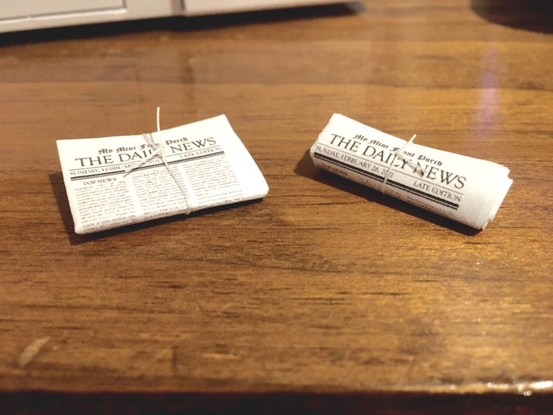 May include: Two rolled up newspapers on a wooden surface. The newspapers are tied with string and have the text "The Daily News" printed on the front.