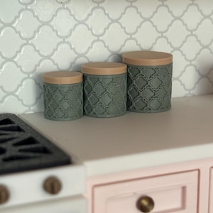 May include: A set of three miniature kitchen canisters in varying sizes. The canisters are a muted green with a textured pattern and tan lids. They are arranged on a white countertop with a white and gray tile backsplash.