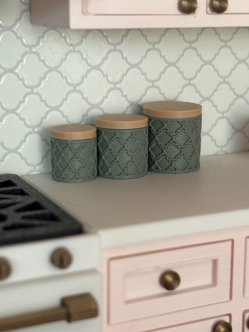 May include: A set of three miniature kitchen canisters in varying sizes. The canisters are a muted green with a textured pattern and tan lids. They are arranged on a white countertop with a white and gray tile backsplash.