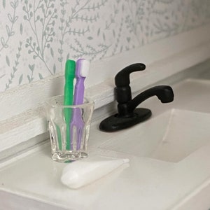 May include: A white bathroom sink with a black tap and a clear glass with two toothbrushes, one green and one purple.