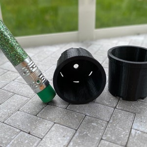 May include: Two black plastic plant pots, one with a drainage hole, on a gray tiled surface. A green glitter pencil is lying on the surface.