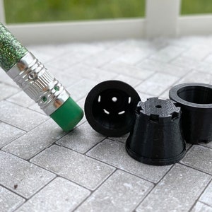 May include: Three small, black, 3D-printed miniature flower pots with varying designs. A green glitter pencil with a silver ferrule and green eraser is positioned near the pots. The pots are on a gray brick-patterned surface.