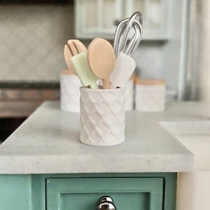 May include: White ceramic utensil holder with a diamond pattern. The holder contains a wooden spoon, a wooden spatula, a green spatula, and a silver whisk.