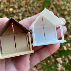 May include: Two miniature wooden houses, one brown and one white, with open sides and a roof. The houses are sitting on a hand.