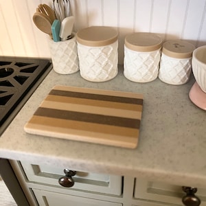 Op de afbeelding: Een wit aanrecht met een bruin en beige gestreept snijplank, vier witte keramische potten met beige deksels en een bestekhouder met houten bestek en een blauwe spatel.