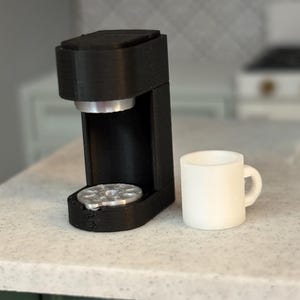 May include: A black single-serve coffee maker with a white ceramic mug on a white countertop.