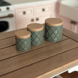 May include: Three decorative canisters in varying sizes. The canisters are olive green with a repeating geometric pattern and tan lids. They are arranged on a wooden table, suggesting a kitchen or home decor theme.