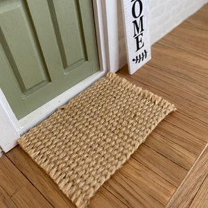 May include: A natural fiber doormat in a light brown color, placed on a wooden floor in front of a green door. The doormat is made of woven fibers and has a rectangular shape.