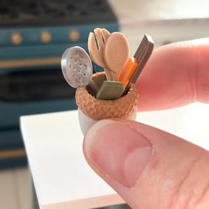 May include: Miniature kitchen utensils in a small, textured basket. The set includes a slotted spoon, a spatula, a fork, and a spoon, along with other small tools. The basket is held in a hand, with a white surface and a teal stove in the background.