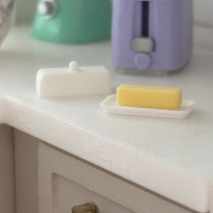 May include: A white ceramic butter dish with a lid and a stick of butter on a white ceramic plate. The butter dish and plate are on a light brown countertop.