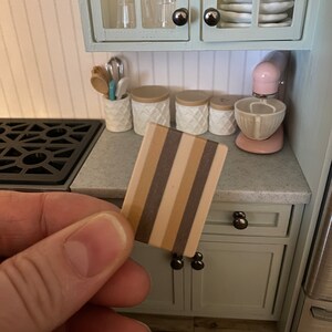 Op de afbeelding: Een miniatuur houten snijplank met bruine en beige strepen. De plank wordt in een hand gehouden voor een poppenhuis keuken.