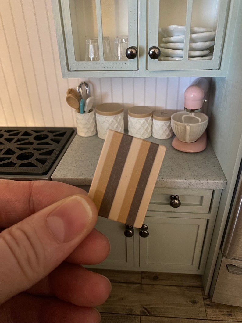 Op de afbeelding: Een miniatuur houten snijplank met bruine en beige strepen. De plank wordt in een hand gehouden voor een poppenhuis keuken.