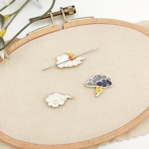 May include: An embroidery hoop with beige fabric displays three cloud-themed enamel pins. One pin shows a sun behind a cloud, another a simple cloud, and the third a storm cloud with a lightning bolt. A needle is inserted through the fabric.