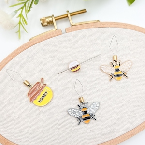 May include: A wooden embroidery hoop holds a canvas with four bee-themed stitch markers. The markers include a honey pot, a bee, and two bee-shaped charms. The markers are yellow, black, and white, with a gold-colored metal finish.