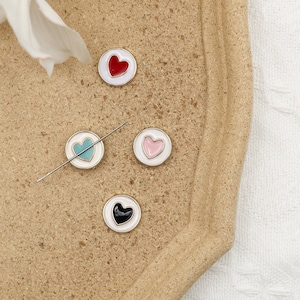 May include: Four heart-shaped buttons with white, red, pink, and black enamel on a beige tray. The buttons are round and have a silver metal rim.