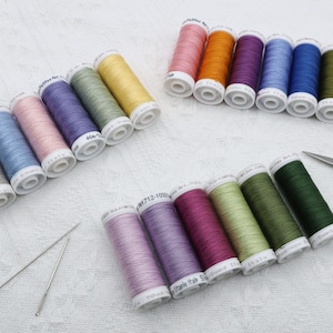 May include: A collection of spools of thread in various colors, including beige, light blue, pink, purple, green, yellow, orange, red, and dark green. The spools are arranged in two groups, with some spools lying on their side and others standing upright. There are also two sewing needles and a pair of scissors in the image.