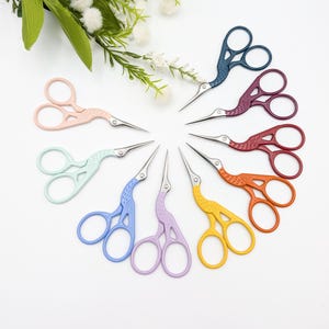 May include: A collection of ten colorful embroidery scissors arranged in a circle. Each scissor features a crane design with a different color, including pink, blue, green, purple, yellow, orange, red, and burgundy. The scissors have silver blades.