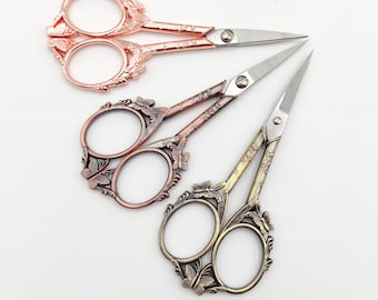 May include: Three pairs of decorative scissors with ornate butterfly and floral designs. The scissors are in rose gold, copper, and silver.