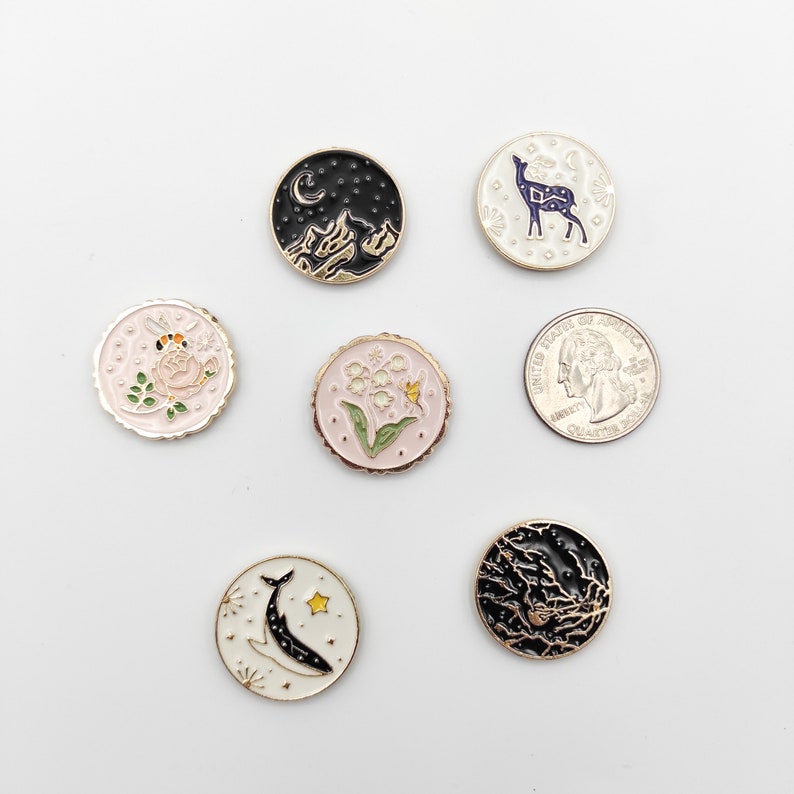May include: A collection of six circular enamel pins featuring various designs including a mountain landscape, a deer, a bee on a flower, lily of the valley, a whale, and a forest scene, displayed next to a US quarter coin for scale reference.