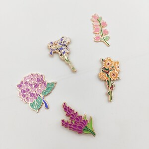 May include: Five enamel pins featuring different floral designs. The pins are gold-toned and feature purple, pink, yellow, and green enamel. The flowers include a hydrangea, a lavender sprig, a cluster of daisies, a single iris, and a sprig of pink flowers.
