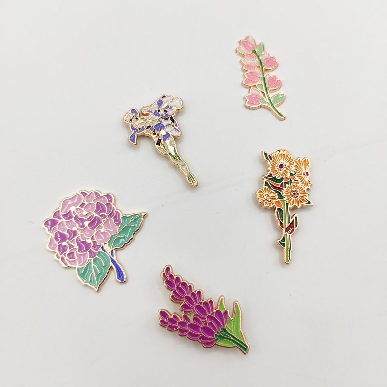 May include: Five enamel pins featuring different floral designs. The pins are gold-toned and feature purple, pink, yellow, and green enamel. The flowers include a hydrangea, a lavender sprig, a cluster of daisies, a single iris, and a sprig of pink flowers.