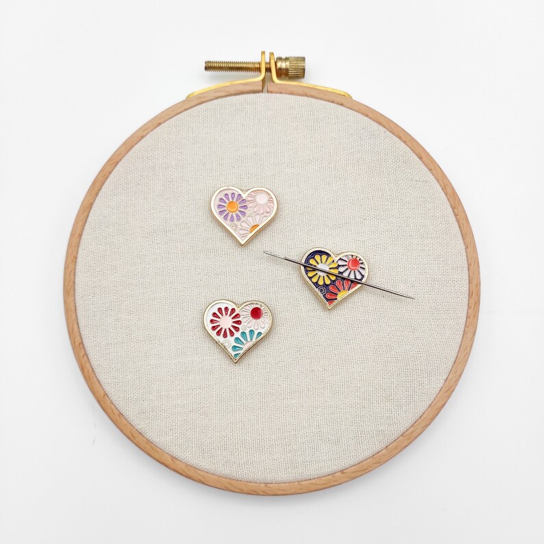 May include: Three gold heart-shaped pins with colorful floral designs on a white fabric background. The pins are displayed on a wooden embroidery hoop.