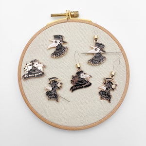May include: Six black and white enamel plague doctor charms and pins arranged on a round wooden embroidery hoop. Some pieces feature a scythe and the text No more germs. Several items include attached metal needle threaders or a sewing needle.