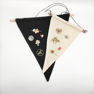 May include: Two black and white triangular fabric pennants with wooden dowels at the top. The pennants are strung together with black and white twine. The black pennant has a variety of pins and brooches attached to it, including a hot air balloon, a star, a flower, and a crown. The white pennant has a bee, a heart, a fish, and a few other pins attached to it.