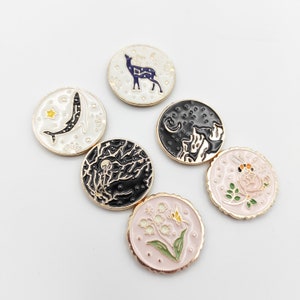 May include: A collection of six circular enamel pins featuring gold-toned edges. Designs include a dark blue deer on white, a white whale on white, a black night sky with mountains, a black tree silhouette, a pink rose with a bee, and lily of the valley.