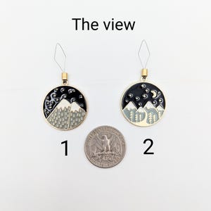 May include: Two round metal charms labelled 1 and 2, each featuring a black night sky with white stars and grey mountains. Charm 2 includes a crescent moon. Both have gold-tone loops and are displayed next to a US quarter for scale. Text reads The view.