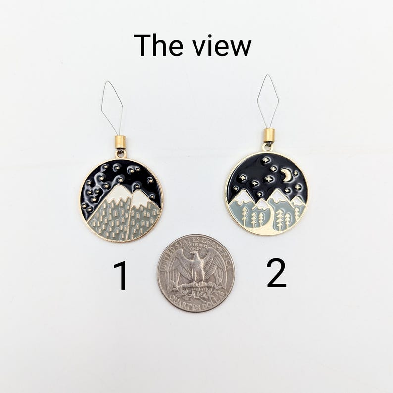 May include: Two round metal charms labelled 1 and 2, each featuring a black night sky with white stars and grey mountains. Charm 2 includes a crescent moon. Both have gold-tone loops and are displayed next to a US quarter for scale. Text reads The view.