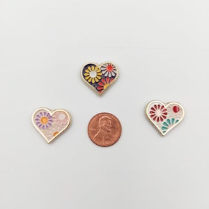 May include: Three gold heart-shaped pins with colorful floral designs. The pins feature different floral patterns in shades of red, yellow, blue, green, and purple.