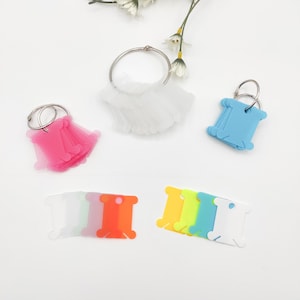 May include: Assortment of colorful plastic thread bobbins with key rings. The bobbins are in various colors, including pink, blue, orange, yellow, and white. There are also clear bobbins with key rings.
