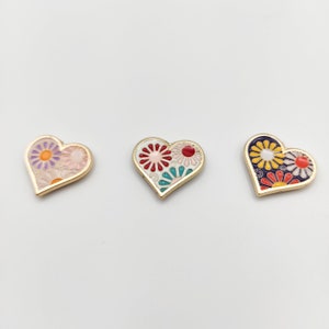May include: Three gold heart-shaped pins with a floral design. The hearts are filled with a pattern of colorful flowers with white, pink, red, yellow, blue, and green accents.