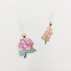 May include: Two gold-toned needle threaders with floral designs. One features a pink and purple hydrangea with green leaves. The other features a pink flower cluster with green leaves.
