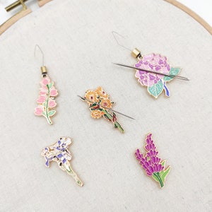 May include: Five needle threaders with different flower designs. The threaders are made of metal and have a gold finish. The flowers are pink, purple, yellow, and white.