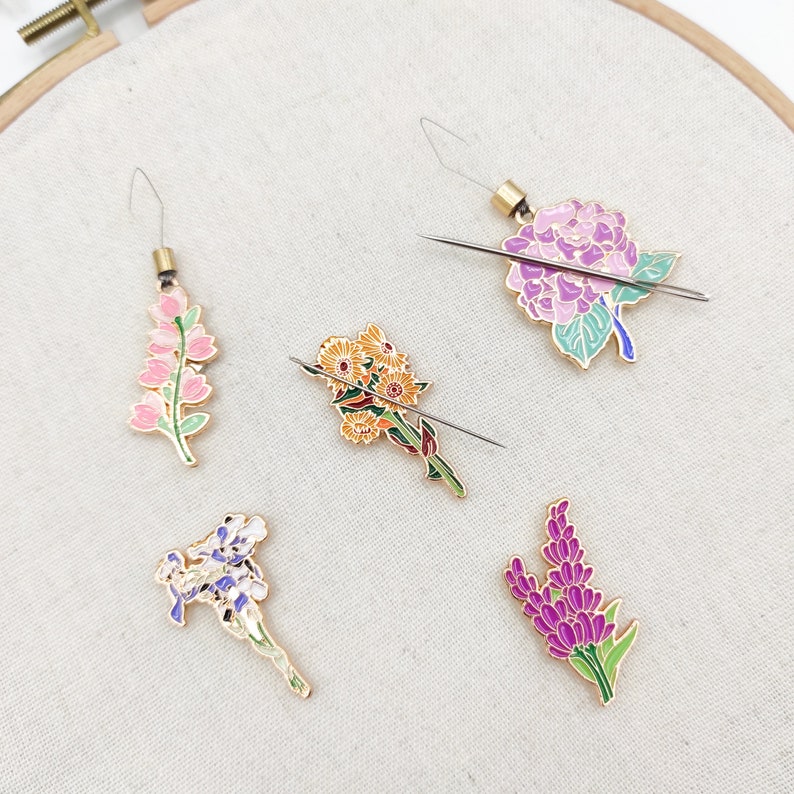 May include: Five needle threaders with different flower designs. The threaders are made of metal and have a gold finish. The flowers are pink, purple, yellow, and white.