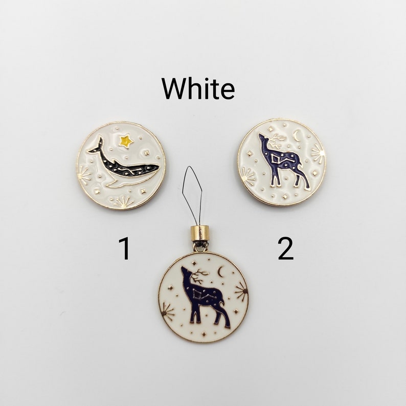 May include: Three round white enamel charms with gold-tone edges. Charm 1 features a dark blue whale and a yellow star. Charms 2 and the bottom charm feature a dark blue deer silhouette with celestial patterns. The bottom charm includes a black loop.