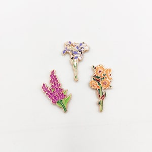 May include: Three enamel pins featuring different floral designs. The first pin is a purple and white flower with a gold outline. The second pin is a purple flower with green leaves and a gold outline. The third pin is a yellow and orange flower with green leaves and a gold outline.