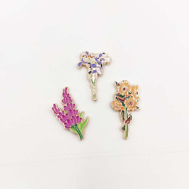 May include: Three enamel pins featuring different floral designs. The first pin is a purple and white flower with a gold outline. The second pin is a purple flower with green leaves and a gold outline. The third pin is a yellow and orange flower with green leaves and a gold outline.
