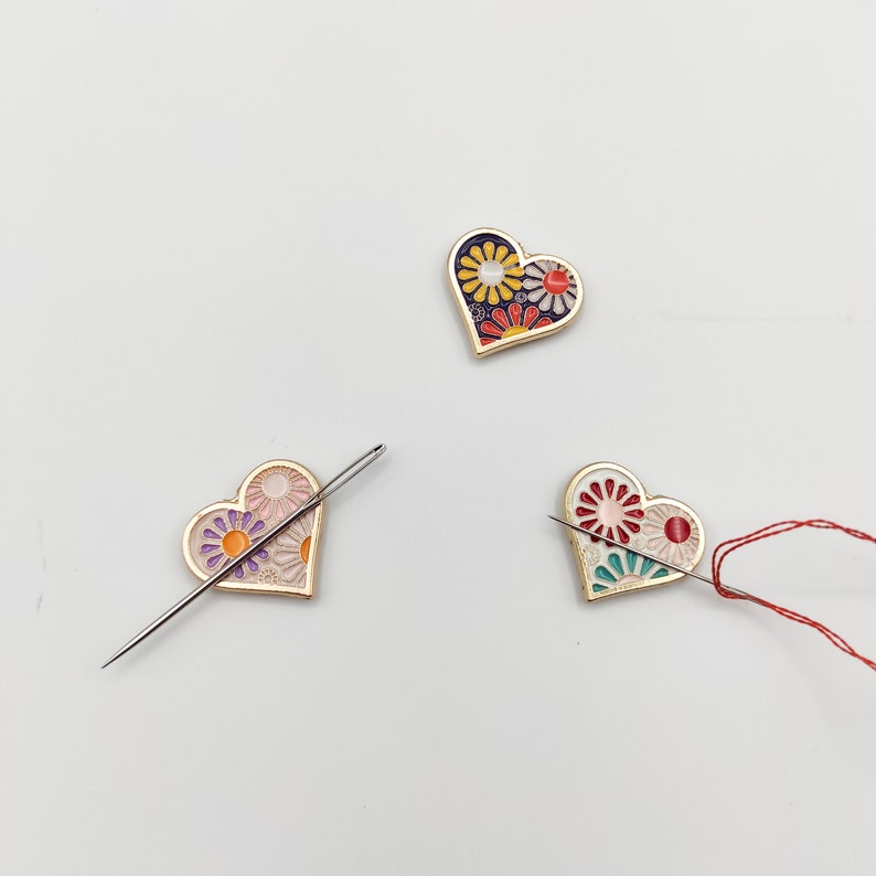 May include: Three heart-shaped pins with floral designs. Each pin has a needle threaded through it. The pins are on a white background.